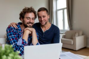 lovely-gay-couple-laughing-together-while-sitting-their-living-room-home-two-romantic-young-male-lovers-having-fun-surfing-internet-indoors-young-gay-couple-livingtogether
